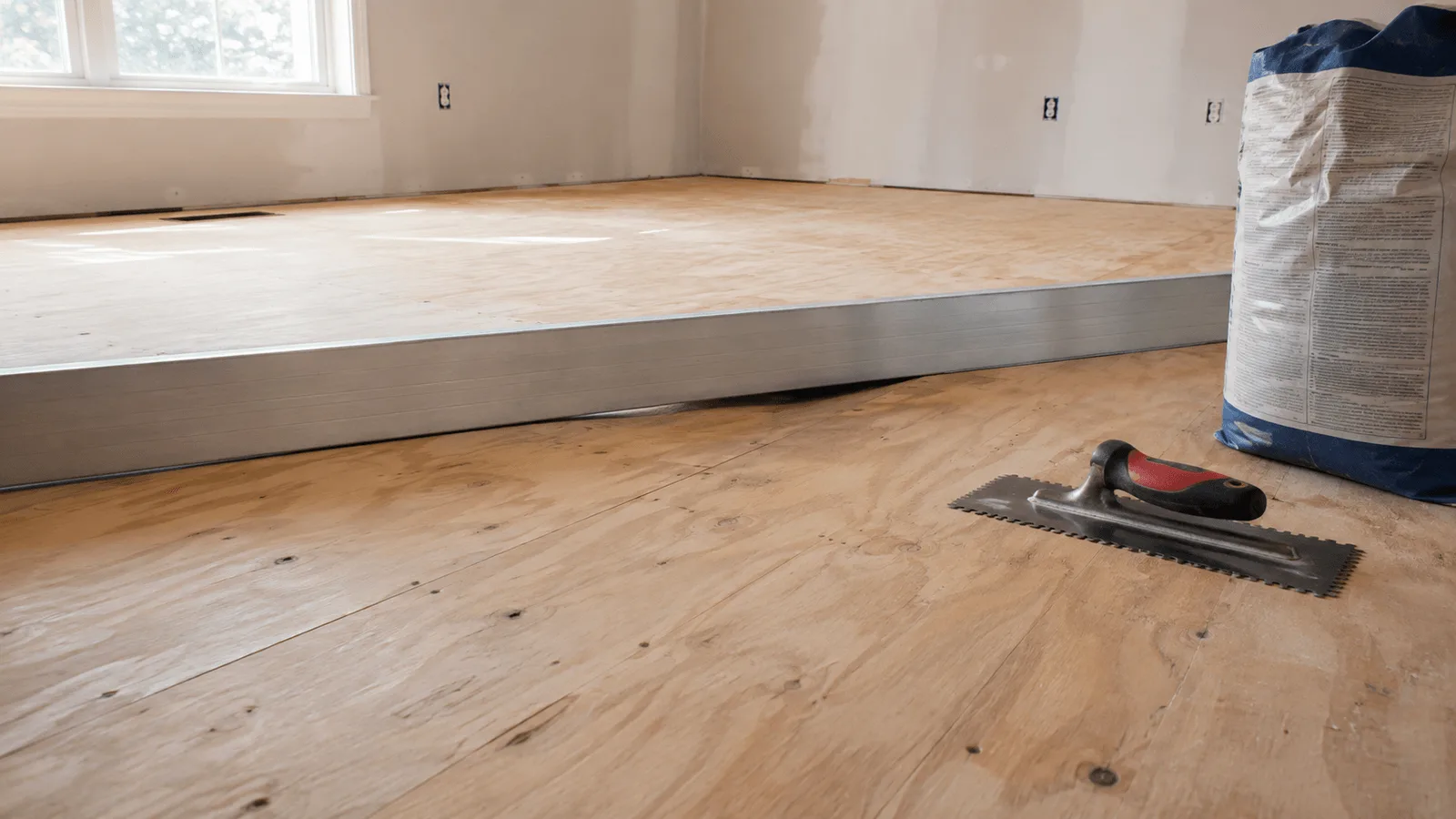 A long straightedge resting across a bare plywood subfloor, showing the gap where the floor dips